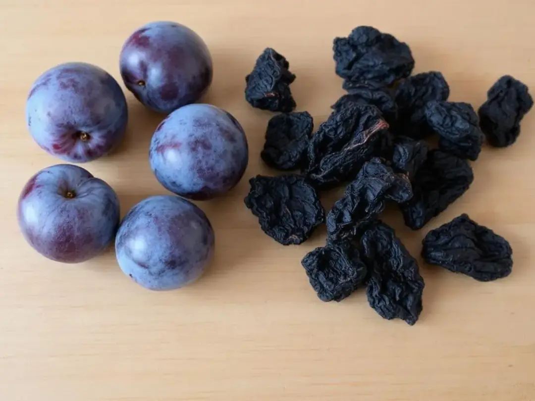 The image shows a comparison between fresh plums on one side and dried prunes on the other, highlighting the difference...