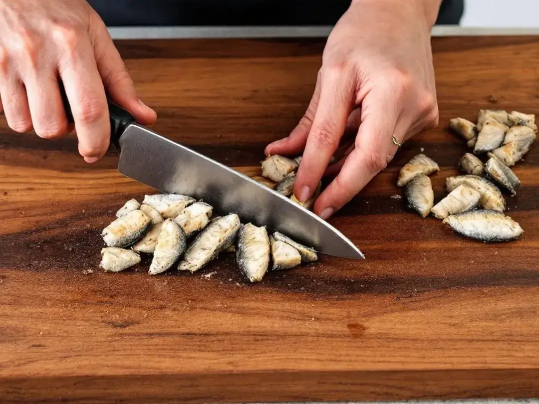 A person's hands are skillfully cutting cooked sardines into small pieces on a wooden cutting board, preparing them for...