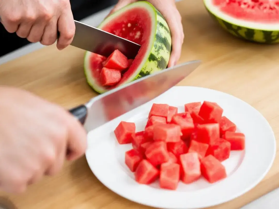 In the image, hands are skillfully cutting a fresh watermelon into bite-sized cubes, ensuring that all seeds and rind...