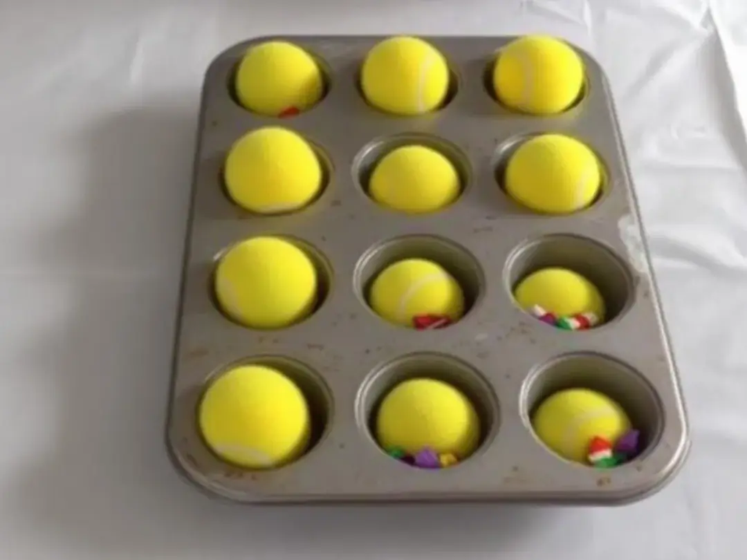 A muffin tin is creatively arranged with tennis balls hiding various tasty treats underneath, serving as an engaging...